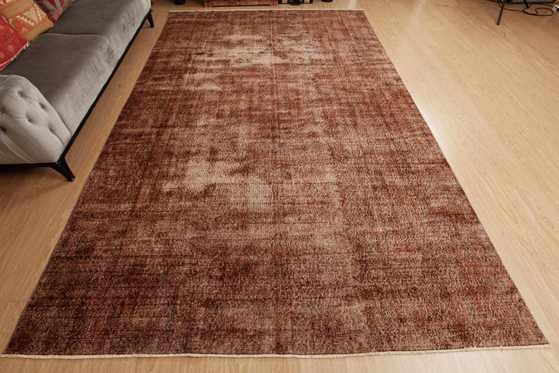 May include: A rectangular area rug featuring a deep brown and terracotta color palette with a distressed, faded texture. The rug displays a subtle, worn-in pattern across its surface, with short fringe detailing visible along the short edges.