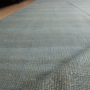 May include: A blue and grey woven rug with a textured surface. The rug is partially rolled up, showing its thickness and construction. The rug is placed on a wooden floor.