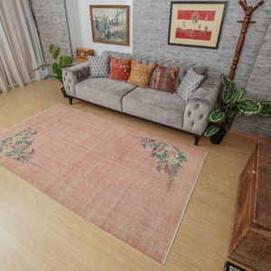 May include: A light pink vintage rug with a floral design. The rug has a faded look and is made of a soft, woven material. The rug is laid out on a hardwood floor in a living room setting.