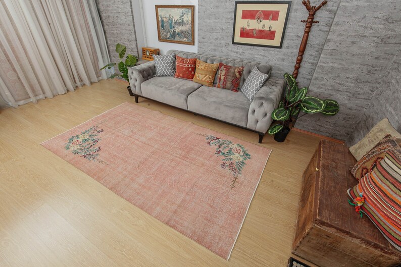 May include: A light pink vintage rug with a floral design. The rug has a faded look and is made of a soft, woven material. The rug is laid out on a hardwood floor in a living room setting.