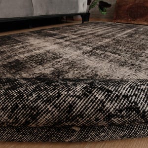 May include: A rectangular area rug featuring a distressed, faded pattern in shades of black, charcoal, and off-white. The rug is partially folded over, revealing a dense, woven texture and a dark, intricate geometric design on its surface.