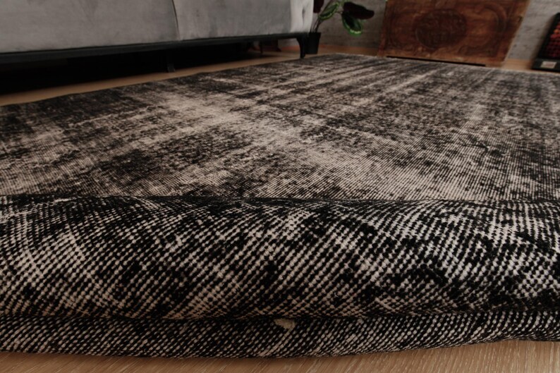 May include: A rectangular area rug featuring a distressed, faded pattern in shades of black, charcoal, and off-white. The rug is partially folded over, revealing a dense, woven texture and a dark, intricate geometric design on its surface.