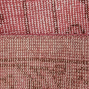 May include: Close-up of a section of a pink and brown rug. The rug has a woven texture with a repeating pattern. The colours are muted, with a mix of pink and brown tones. The rug appears to be made of natural fibres.