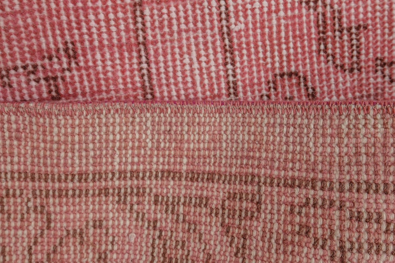 May include: Close-up of a section of a pink and brown rug. The rug has a woven texture with a repeating pattern. The colours are muted, with a mix of pink and brown tones. The rug appears to be made of natural fibres.