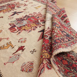 Puede incluir: Una alfombra de color crema con un patrón geométrico rojo, azul y amarillo. La alfombra tiene un borde de flecos y está doblada por la mitad.