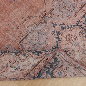 May include: A woven rug featuring a faded terracotta and dusty rose base with intricate dark blue floral and scrollwork patterns. The textile displays a distressed texture with a corner folded over to reveal the underside and detailed weave structure.