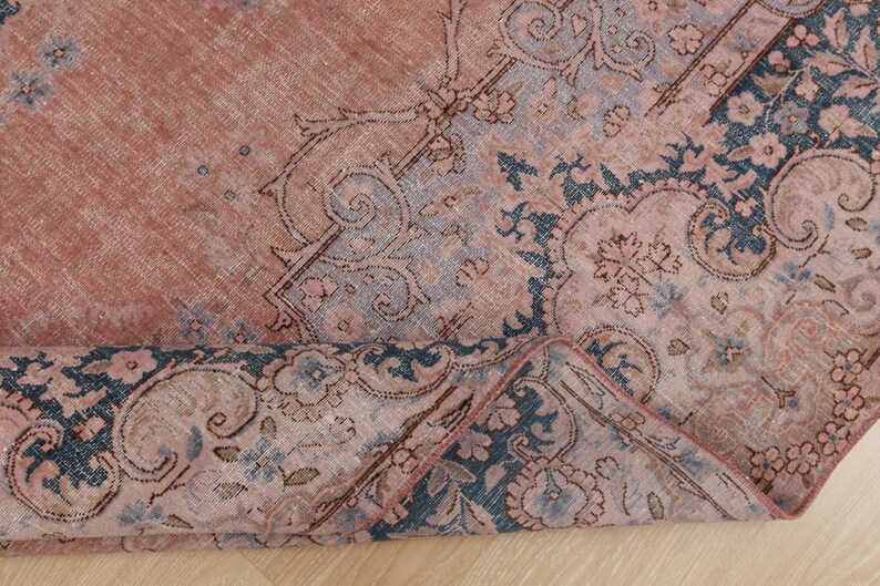 May include: A woven rug featuring a faded terracotta and dusty rose base with intricate dark blue floral and scrollwork patterns. The textile displays a distressed texture with a corner folded over to reveal the underside and detailed weave structure.
