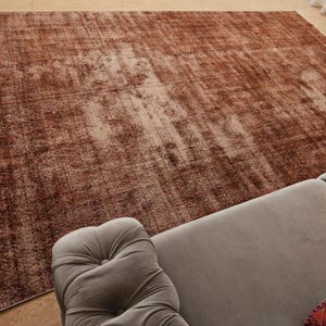 May include: A large rectangular area rug featuring a distressed, mottled pattern in shades of deep rust, copper, and faded brown. The rug has a low-pile texture with visible vertical lines and a light-colored fringe along the left edge.