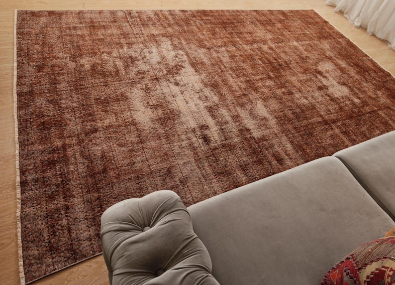 May include: A large rectangular area rug featuring a distressed, mottled pattern in shades of deep rust, copper, and faded brown. The rug has a low-pile texture with visible vertical lines and a light-colored fringe along the left edge.
