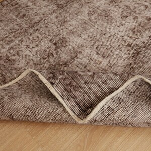 May include: A section of a brown and beige patterned rug, with a folded edge revealing the underside. The rug has a distressed, vintage appearance, with a faded floral design. The rug is placed on a light-colored wooden surface.