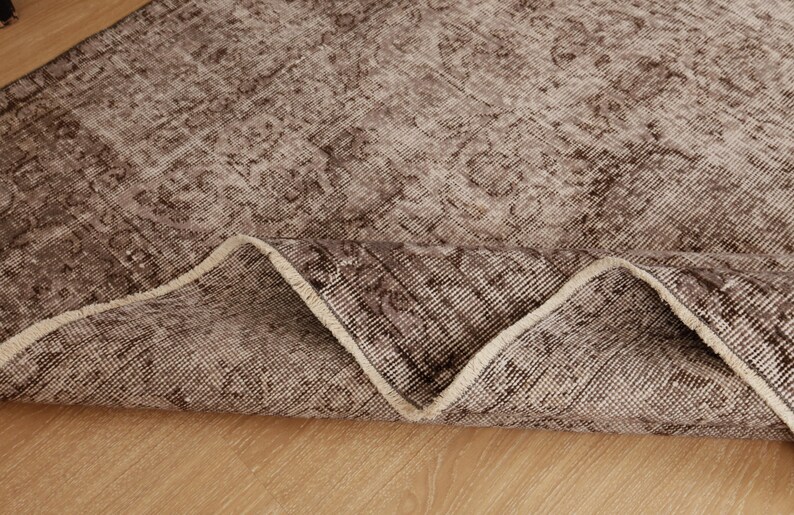 May include: A section of a brown and beige patterned rug, with a folded edge revealing the underside. The rug has a distressed, vintage appearance, with a faded floral design. The rug is placed on a light-colored wooden surface.