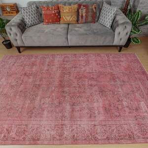 May include: A large, rectangular rug in shades of pink and faded black, with a distressed appearance. The rug has an ornate border and a central design with geometric and floral elements. It is placed on a wooden floor.