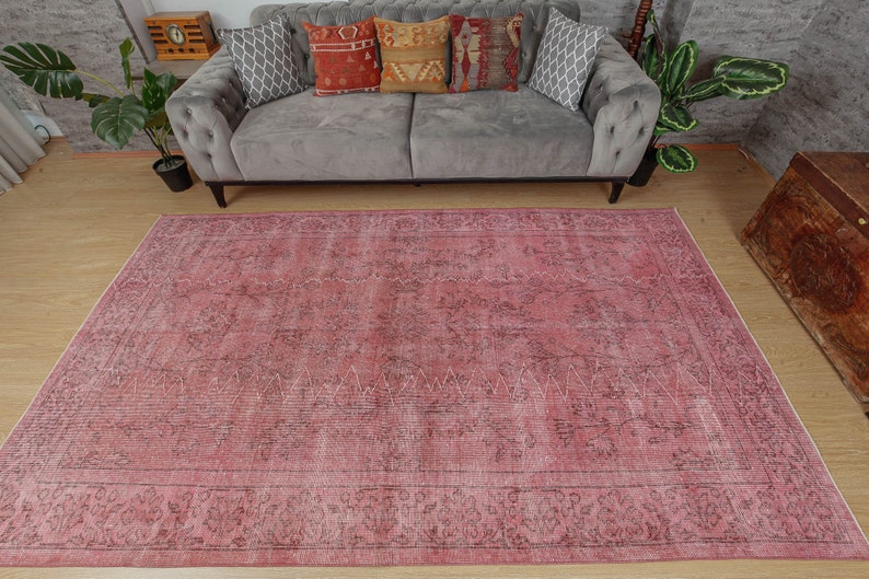 May include: A large, rectangular rug in shades of pink and faded black, with a distressed appearance. The rug has an ornate border and a central design with geometric and floral elements. It is placed on a wooden floor.