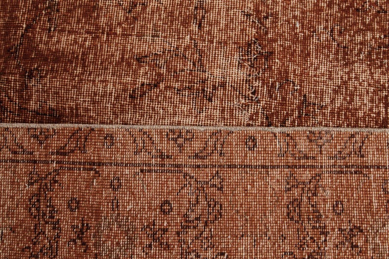 May include: A woven rug featuring a deep rust-brown color palette with a distressed, weathered texture. The surface displays a repeating pattern of dark brown linear motifs and geometric shapes arranged in a structured border and field design.
