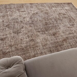 May include: A large, rectangular rug with a distressed, vintage appearance. The rug features a blend of brown and beige tones, creating a textured, mottled effect. The rug is placed on a light-colored wooden floor, with a gray sofa in the foreground.