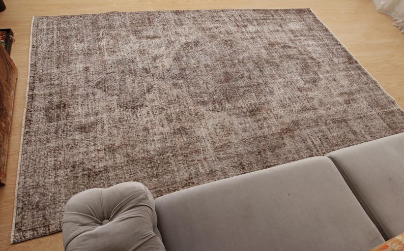 May include: A large, rectangular rug with a distressed, vintage appearance. The rug features a blend of brown and beige tones, creating a textured, mottled effect. The rug is placed on a light-colored wooden floor, with a gray sofa in the foreground.