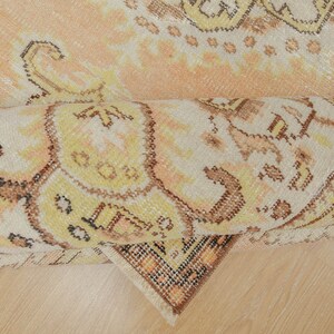 May include: Rolled-up vintage rug with a faded, distressed appearance. The rug features a complex, ornate pattern in shades of peach, cream, yellow, and brown. The design includes floral and geometric elements, creating a traditional aesthetic.
