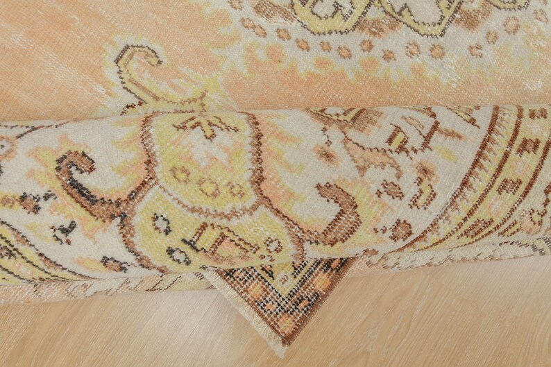 May include: Rolled-up vintage rug with a faded, distressed appearance. The rug features a complex, ornate pattern in shades of peach, cream, yellow, and brown. The design includes floral and geometric elements, creating a traditional aesthetic.