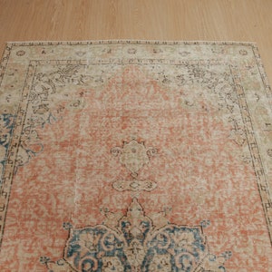 Puede incluir: Una alfombra vintage con un fondo melocot&oacute;n descolorido y un patr&oacute;n floral azul y beige. La alfombra tiene un aspecto desgastado y descolorido y un borde con flecos.