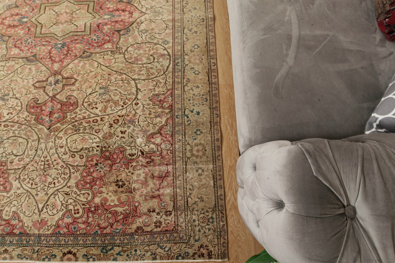 Pu&ograve; includere: Un tappeto in stile antico con un motivo floreale dettagliato nei toni del beige, rosso e blu. Il design del tappeto include un medaglione centrale e bordi ornati. Una sezione di un divano grigio imbottito &egrave; visibile a destra.