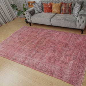 May include: A large, rectangular rug in shades of pink and burgundy with a distressed appearance. The rug has an ornate border and a faded, vintage-style pattern. It's placed on a light wooden floor, with a grey sofa and decorative cushions in the background.