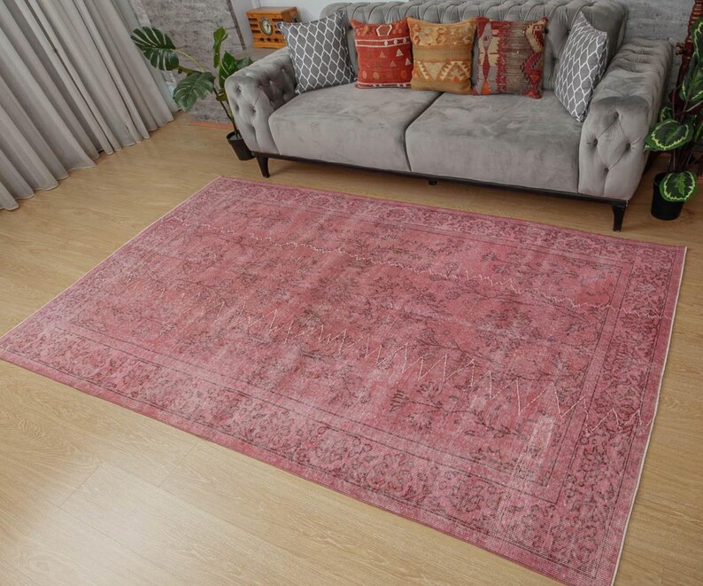 May include: A large, rectangular rug in shades of pink and burgundy with a distressed appearance. The rug has an ornate border and a faded, vintage-style pattern. It's placed on a light wooden floor, with a grey sofa and decorative cushions in the background.