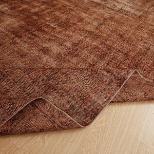 May include: A large area rug featuring a deep rust and brown color palette with a distressed, faded pattern. The rug is folded over at one corner, revealing the textured weave and the underside of the fabric, showcasing a consistent tonal design.
