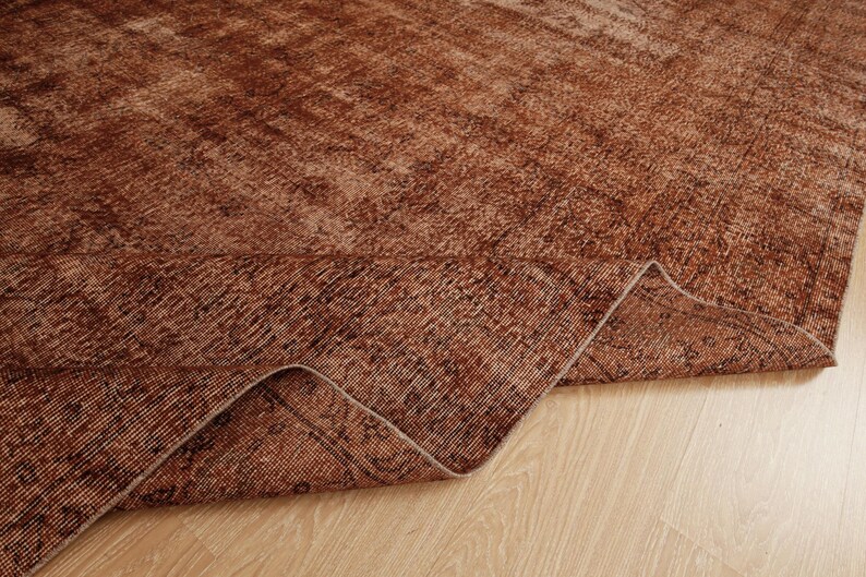 May include: A large area rug featuring a deep rust and brown color palette with a distressed, faded pattern. The rug is folded over at one corner, revealing the textured weave and the underside of the fabric, showcasing a consistent tonal design.