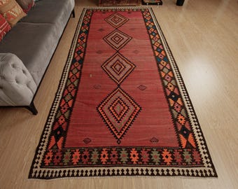 May include: A red, black, and orange patterned rug with a diamond design. The rug has a fringe border and is laid out on a wooden floor.