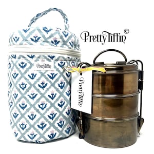 May include: A patterned lunch bag and a three-tiered metal tiffin carrier. The bag features a blue and white floral design and a handle. The bronze-coloured tiffin carrier has a handle on top. The brand name "Pretty Tiffin" is visible.