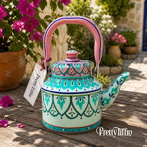 May include: A hand-painted teapot with a pink handle and lid. The teapot features a teal and pink floral design with the text "Pretty Tiffin" on a tag and on the teapot itself. The teapot is sitting on a wooden surface.
