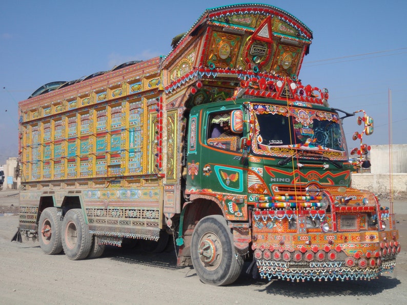 May include: A brightly decorated green HINO truck with intricate painted designs and colorful embellishments. The truck has a large cargo bed with multiple compartments and is adorned with numerous lights and ornaments.