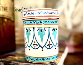 May include: A cylindrical, white ceramic container with a lid, adorned with teal, orange, and blue floral patterns. The design includes stylized leaves and geometric shapes. The container is likely for storage or decoration.