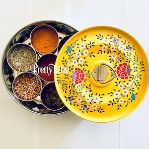Puede incluir: Un recipiente para especias de metal amarillo con un diseño floral. La tapa tiene un patrón floral con acentos rojos, blancos y azules. El recipiente tiene seis compartimentos para especias.