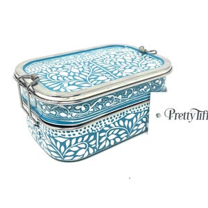 Puede incluir: Una fiambrera de acero inoxidable de dos niveles con un diseño floral en color turquesa y blanco. La fiambrera tiene un cierre y un borde de metal plateado. Las palabras "Pretty Tiffin" están en la esquina inferior derecha.