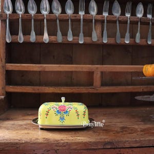 Op de afbeelding: Gele botervloot met een bloemenpatroon. De vloot heeft een zilveren deksel en staat op een houten oppervlak.