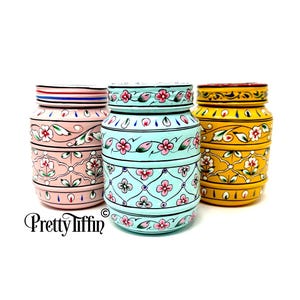 May include: Three decorative ceramic jars in pink, turquoise, and yellow. Each jar features a floral design with a lid. The pink jar has a red, white, and blue striped band. The jars are labeled "Pretty Tiffin".