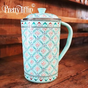 May include: A turquoise blue ceramic jug with a floral pattern and a silver lid. The jug has a handle and is decorated with red and green accents.