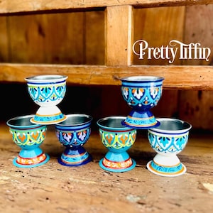 May include: A set of six colorful egg cups with a stainless steel interior. The cups feature a variety of designs in blue, orange, and white, with the text "Pretty Tiffin" in the upper right corner. The cups are displayed on a wooden surface.
