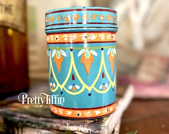 May include: A hand-painted teal jar with a lid, featuring orange, yellow, and white floral patterns. The jar has the text "Pretty Tiffin" on the side. The jar is sitting on top of a book.