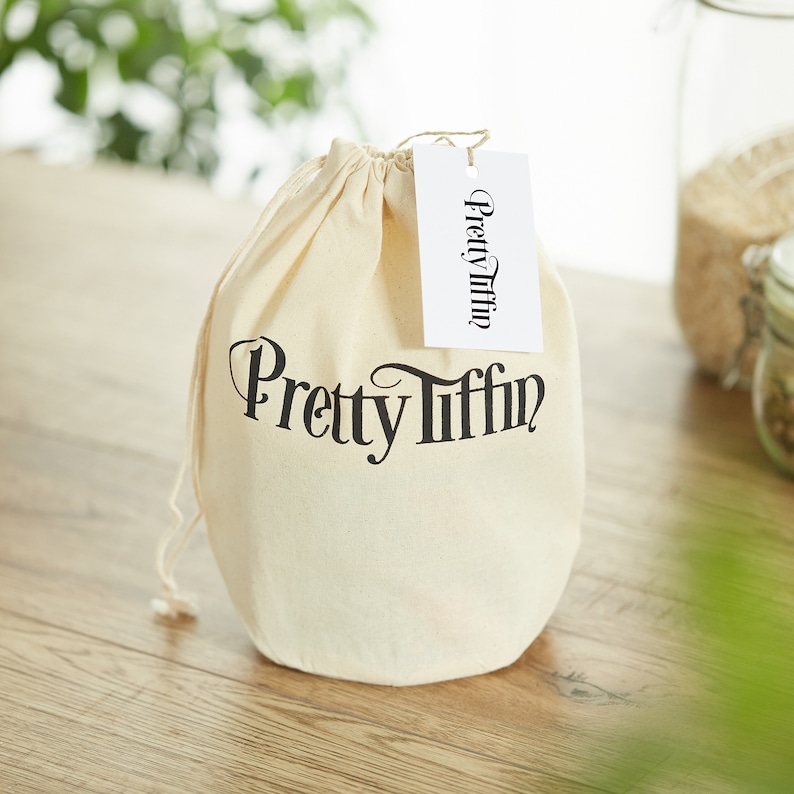 May include: A white cotton drawstring bag with the text "Pretty Tiffin" printed in black on the front. The bag is sitting on a wooden surface.