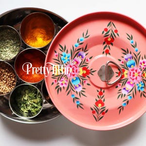 May include: A round, stainless steel spice container with six individual compartments. The container has a pink lid with a floral design and a silver handle.