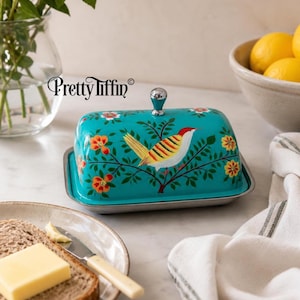 Op de afbeelding: Een rechthoekige turquoise botervloot met een zilverkleurige metalen basis en een bijpassend deksel met een handgeschilderde geel-rode vogel op een tak met oranje en gele bloemen. Het deksel heeft een zilveren knop en de tekst Pretty Tiffin.