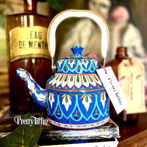 Könnte beinhalten: Eine blau-weiß dekorierte Teekanne mit weißem Griff und passendem Deckel. Die Teekanne hat ein detailliertes Muster mit weißen, orangefarbenen und grünen Akzenten. Das Etikett lautet "Pretty Tiffin?"