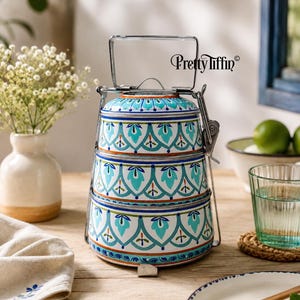 Könnte beinhalten: Eine dreistufige Tiffin-Lunchbox aus Metall mit silbernem Griff und Verschlussmechanismus. Die Außenseite hat eine weiße Basis mit einem sich wiederholenden Muster aus türkisfarbenen, blauen und orangefarbenen Blumenmotiven. Der Text Pretty Tiffin ist in Schwarz aufgedruckt.