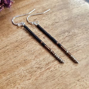 May include: A pair of long, silver dangle earrings with black and copper beads. The earrings are made with small, round beads that are strung together to create a long, thin strand.