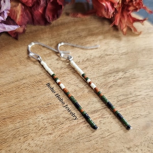 May include: A pair of long, beaded earrings with a white, black, green, and brown color scheme. The earrings are hanging from silver hooks. The text "Boho Hobo Jewelry" is visible in the image.