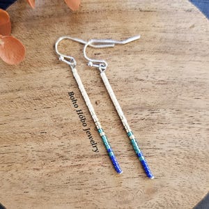 May include: A pair of long, silver dangle earrings with blue, green, and white beads. The earrings are on a wooden surface with the text "boho hobbo jewelry" printed on the surface.