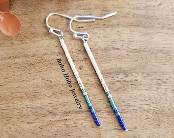 May include: A pair of long, silver dangle earrings with blue, green, and white beads. The earrings are on a wooden surface with the text "boho hobbo jewelry" printed on the surface.
