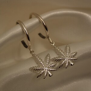 May include: Silver hoop earrings with dangling marijuana leaf charms. The earrings are made of a shiny, metallic material and are displayed on a white satin fabric. The earrings are a unique accessory.
